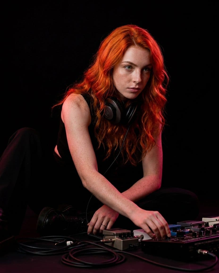 Celeste assise au sol en studio, casque autour du cou, entourée de câbles et de pédales.